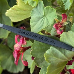 May include: A black object with the text "MADE IN U.S.A." is set against a background of green leaves and red flowers. The leaves are a bright green, and the flowers are a deep red.