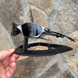 以下が含まれることがあります： 黒いスタンドに展示された、ダークレンズのシルバーフレームのOakleyサングラス。スタンドにはOakleyのロゴがあり、石壁を背景に手で持たれています。 サングラスは洗練されたモダンなデザインです。
