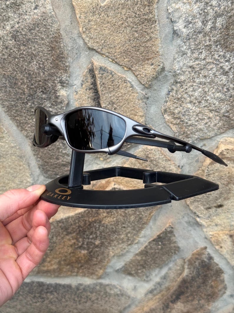 以下が含まれることがあります： 黒いスタンドに展示された、ダークレンズのシルバーフレームのOakleyサングラス。スタンドにはOakleyのロゴがあり、石壁を背景に手で持たれています。 サングラスは洗練されたモダンなデザインです。