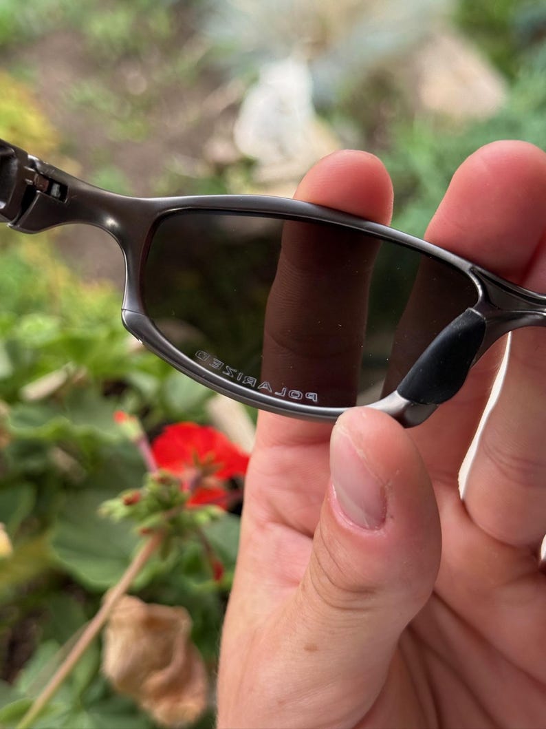 May include: Close-up of a pair of gray sunglasses with dark lenses. The sunglasses have a sleek, modern design with a metal frame and black nose pads. The word "POLARIZED" is printed on the lens.