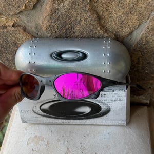 May include: Black Oakley sunglasses with bright pink mirrored lenses. The sunglasses are held in front of a silver case and a white box with black text. The lenses reflect the surrounding environment.