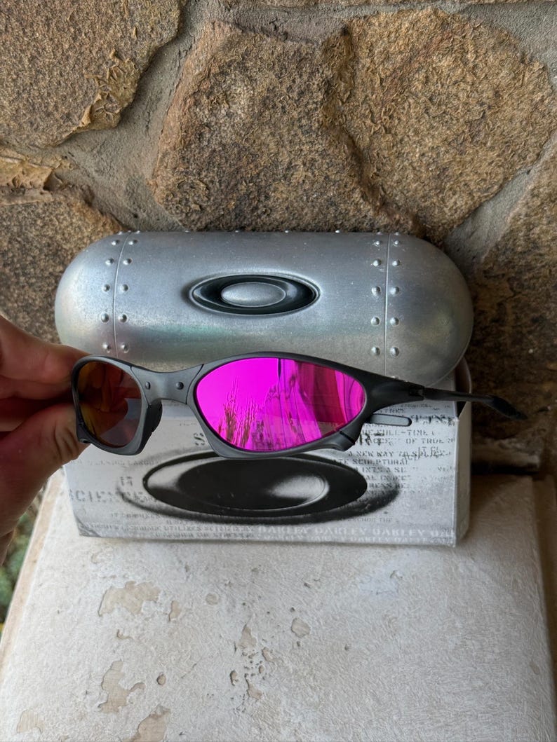 May include: Black Oakley sunglasses with bright pink mirrored lenses. The sunglasses are held in front of a silver case and a white box with black text. The lenses reflect the surrounding environment.