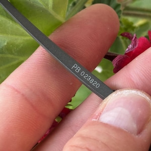 May include: Close-up of a dark grey metal object with the text "PB 023820" etched on its surface. The object is held between fingers, with a blurred background of green leaves and red flowers.