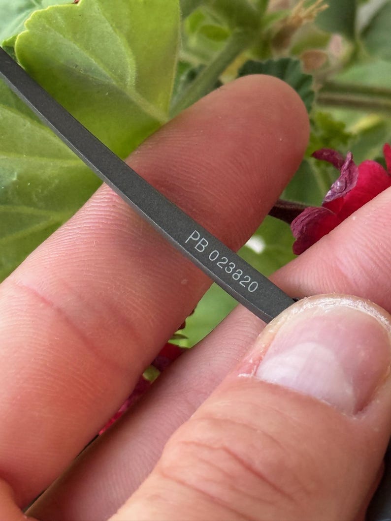May include: Close-up of a dark grey metal object with the text "PB 023820" etched on its surface. The object is held between fingers, with a blurred background of green leaves and red flowers.