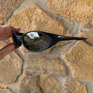 K&ouml;nnte beinhalten: Schwarze Sonnenbrille mit ovalen Gl&auml;sern und einem geschwungenen Rahmen. Die Sonnenbrille hat ein schlankes, modernes Design und wird vor einem Hintergrund aus strukturiertem Stein gehalten.