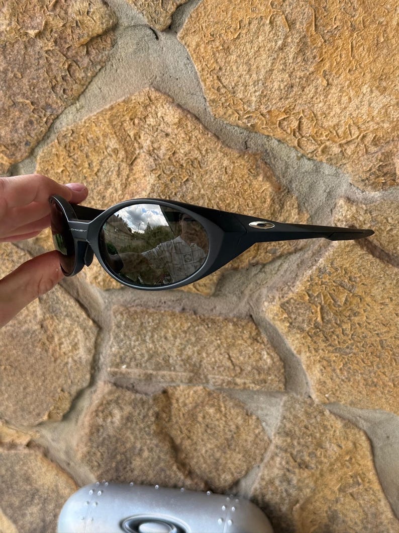 K&ouml;nnte beinhalten: Schwarze Sonnenbrille mit ovalen Gl&auml;sern und einem geschwungenen Rahmen. Die Sonnenbrille hat ein schlankes, modernes Design und wird vor einem Hintergrund aus strukturiertem Stein gehalten.