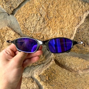 May include: Black sunglasses with purple lenses. The sunglasses have a sleek, modern design with a thin frame and a reflective lens. The lenses are a vibrant purple color, and the frame is black.