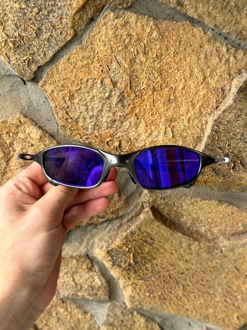 May include: Black sunglasses with purple lenses. The sunglasses have a sleek, modern design with a thin frame and a reflective lens. The lenses are a vibrant purple color, and the frame is black.