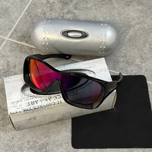 May include: Black Oakley sunglasses with red and purple mirrored lenses, resting on a white box with text and a black cleaning cloth. A silver case with the Oakley logo is in the background.