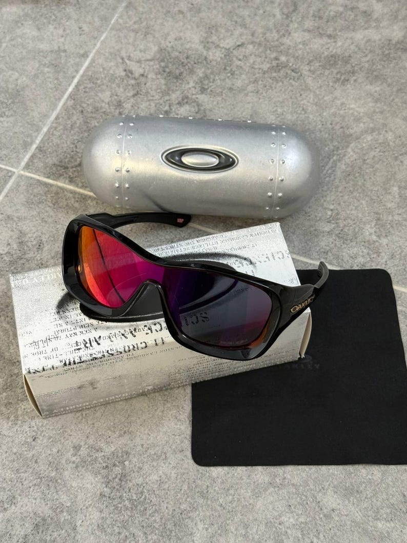 May include: Black Oakley sunglasses with red and purple mirrored lenses, resting on a white box with text and a black cleaning cloth. A silver case with the Oakley logo is in the background.