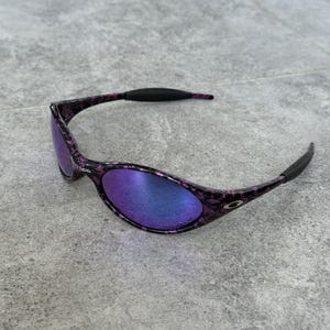 Puede incluir: Gafas de sol ovaladas con montura moteada morada y negra y lentes espejadas que reflejan tonos azules y morados. Las patillas son negras y curvadas. Las lentes son ligeramente curvadas.