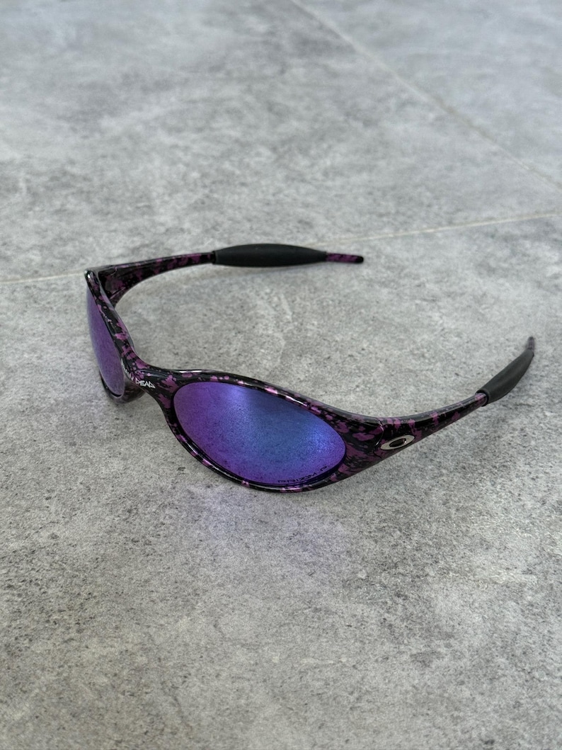 Puede incluir: Gafas de sol ovaladas con montura moteada morada y negra y lentes espejadas que reflejan tonos azules y morados. Las patillas son negras y curvadas. Las lentes son ligeramente curvadas.