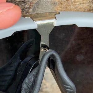 May include: Close-up of gray and black sunglasses. The sunglasses have a silver metal bridge and black nose pads. The lenses are dark, and the frame is a light gray color. The sunglasses are partially obscuring the view.
