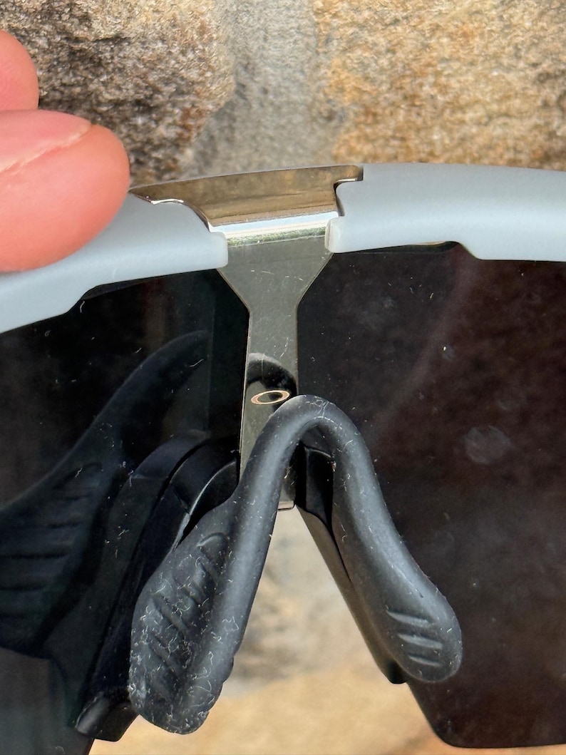 May include: Close-up of gray and black sunglasses. The sunglasses have a silver metal bridge and black nose pads. The lenses are dark, and the frame is a light gray color. The sunglasses are partially obscuring the view.