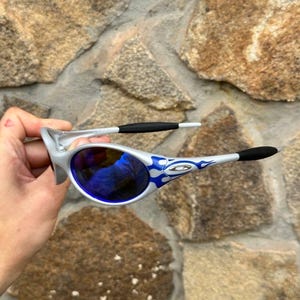 May include: Silver-framed sunglasses with blue lenses and blue flame designs on the arms. The sunglasses have black earpieces and a silver bridge. The lenses reflect the surrounding environment. The sunglasses are held against a stone wall background.