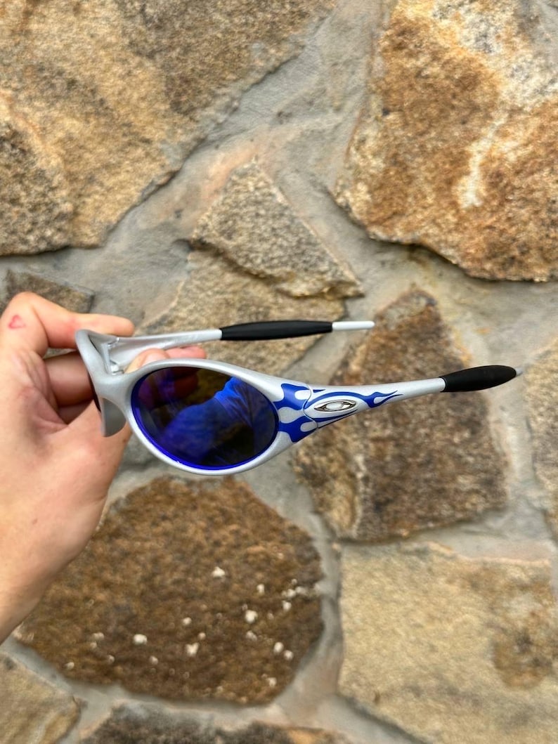 May include: Silver-framed sunglasses with blue lenses and blue flame designs on the arms. The sunglasses have black earpieces and a silver bridge. The lenses reflect the surrounding environment. The sunglasses are held against a stone wall background.