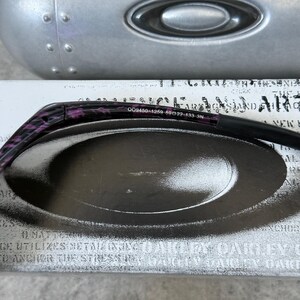 Puede incluir: Gafas de sol con estampado negro y morado con el texto "009450-1259 59[]22-133 3N" en la patilla. Las gafas de sol est&aacute;n sobre un estuche plateado con un logotipo ovalado negro.