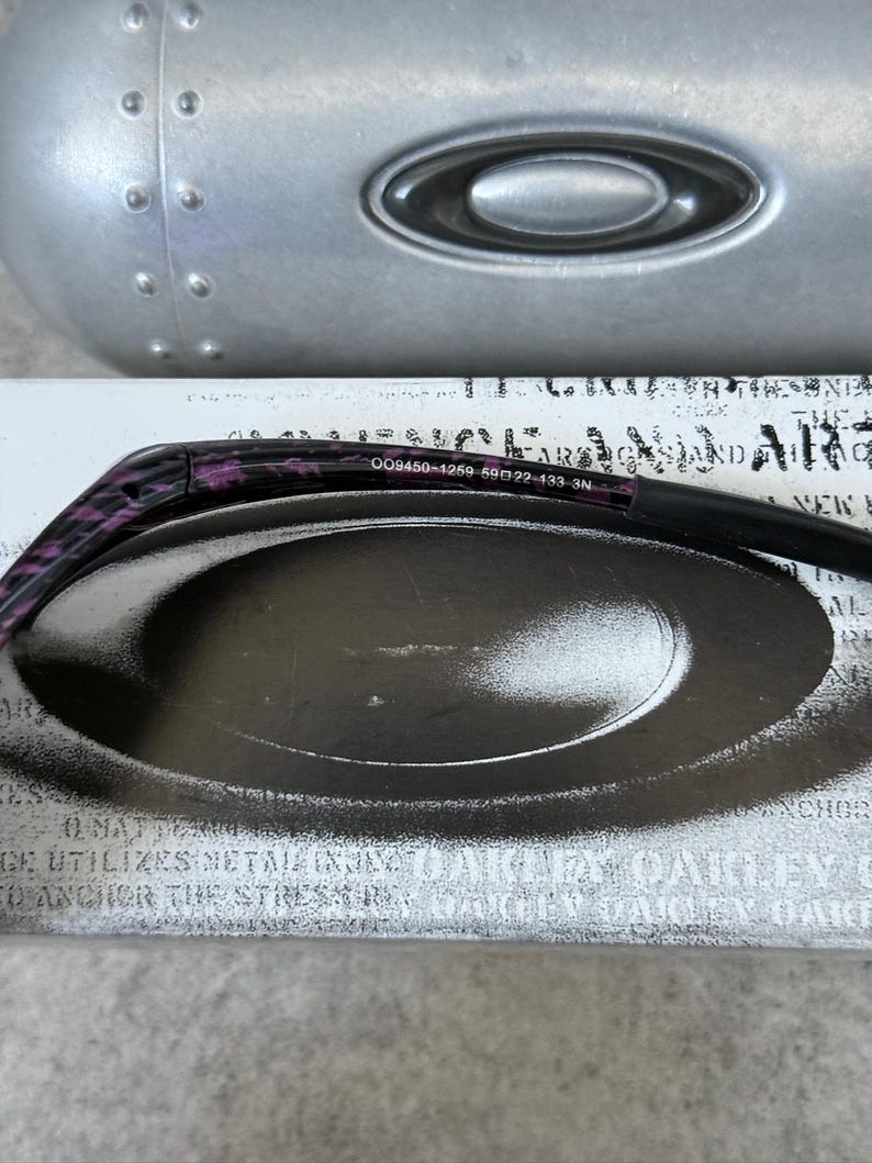 Puede incluir: Gafas de sol con estampado negro y morado con el texto "009450-1259 59[]22-133 3N" en la patilla. Las gafas de sol est&aacute;n sobre un estuche plateado con un logotipo ovalado negro.