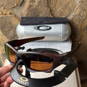 Może przedstawiać: Ciemnobrązowe okulary przeciwsłoneczne Oakley z pomarańczowymi szkłami, prezentowane na czarnym stojaku ze złotym logo Oakley. Srebrne etui i czarna ściereczka w tle. Sportowy design.