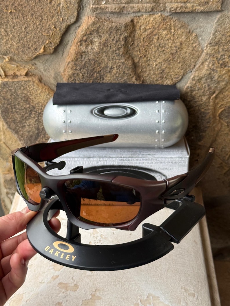 Może przedstawiać: Ciemnobrązowe okulary przeciwsłoneczne Oakley z pomarańczowymi szkłami, prezentowane na czarnym stojaku ze złotym logo Oakley. Srebrne etui i czarna ściereczka w tle. Sportowy design.