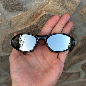 May include: Black sunglasses with mirrored lenses are held in a hand. The lenses reflect the sky and surrounding environment. The sunglasses have a sleek, sporty design, suitable for outdoor activities.