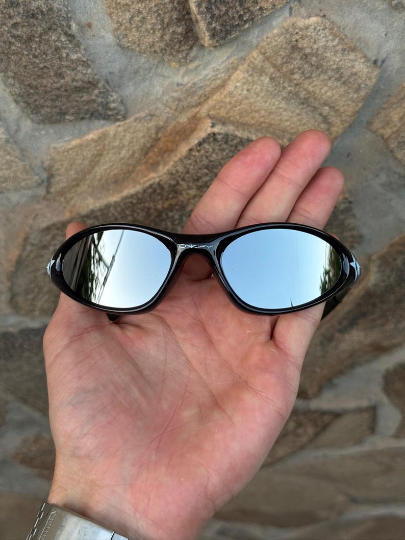 May include: Black sunglasses with mirrored lenses are held in a hand. The lenses reflect the sky and surrounding environment. The sunglasses have a sleek, sporty design, suitable for outdoor activities.