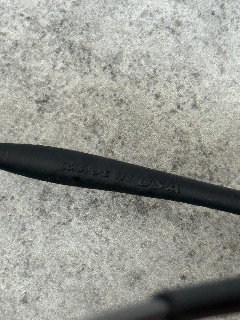 May include: Close-up of a black object with the embossed text "MADE IN USA". The object appears to be a part of a larger item, possibly eyewear or a similar product. The background is a textured gray surface.
