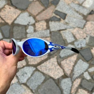 May include: White and blue sunglasses with blue mirrored lenses. The temples feature a blue and white flame design. The sunglasses are held against a stone background.
