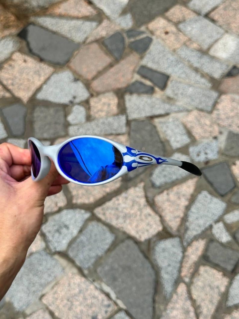 May include: White and blue sunglasses with blue mirrored lenses. The temples feature a blue and white flame design. The sunglasses are held against a stone background.