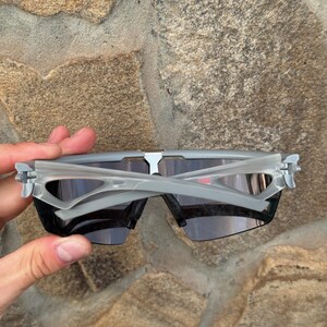 May include: Gray sports sunglasses with dark lenses. The sunglasses have a modern, angular design with a gray frame and temples. The lenses are a dark, smoky color, providing protection from the sun.