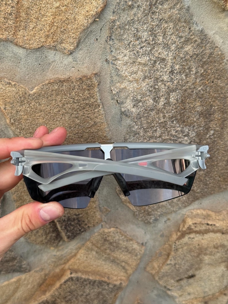 May include: Gray sports sunglasses with dark lenses. The sunglasses have a modern, angular design with a gray frame and temples. The lenses are a dark, smoky color, providing protection from the sun.