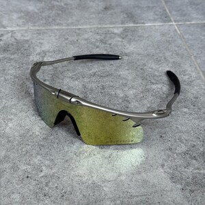 May include: Silver-framed sunglasses with iridescent lenses. The sunglasses have a sleek, modern design with black ear pieces. The lenses have a gradient of yellow and green hues. The sunglasses are resting on a gray surface.