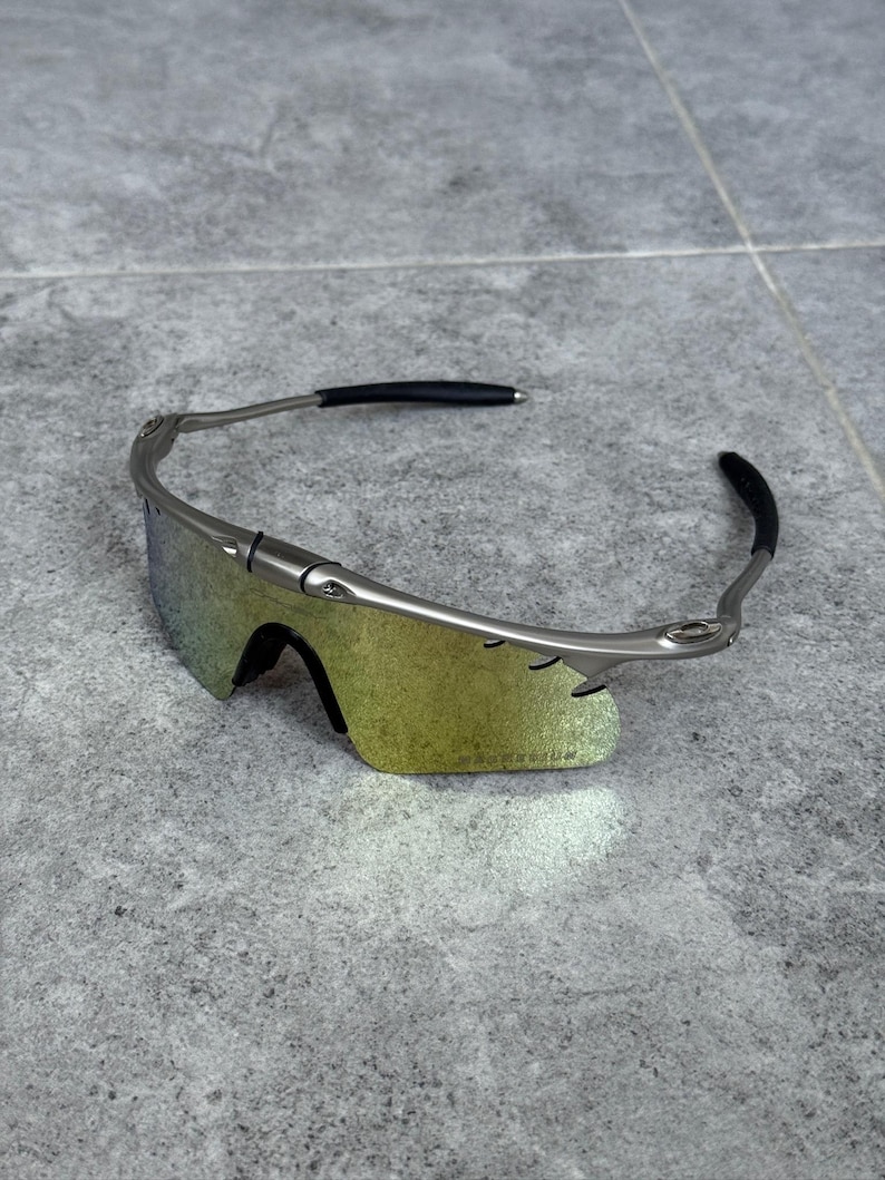 May include: Silver-framed sunglasses with iridescent lenses. The sunglasses have a sleek, modern design with black ear pieces. The lenses have a gradient of yellow and green hues. The sunglasses are resting on a gray surface.