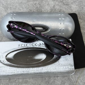 Puede incluir: Gafas de sol negras con un estampado floral p&uacute;rpura en las patillas, sobre un estuche plateado y una caja blanca con la palabra "SCIENCE" impresa. Las gafas tienen lentes oscuras y el logotipo de Oakley es visible.