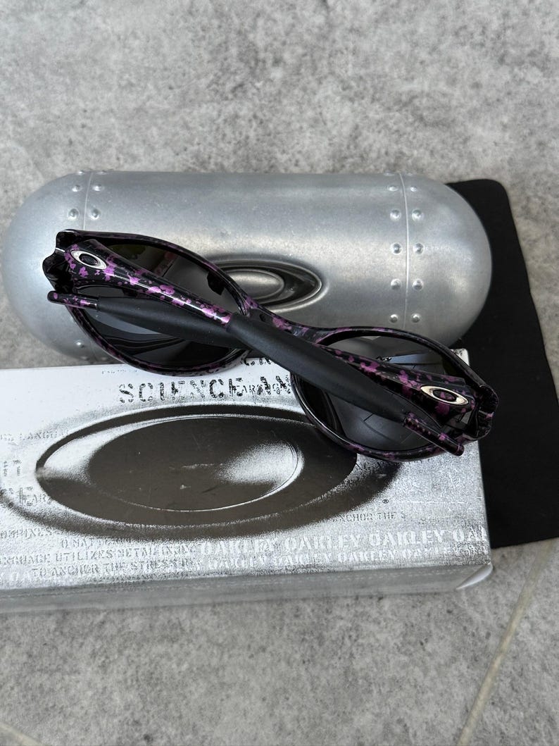 Puede incluir: Gafas de sol negras con un estampado floral p&uacute;rpura en las patillas, sobre un estuche plateado y una caja blanca con la palabra "SCIENCE" impresa. Las gafas tienen lentes oscuras y el logotipo de Oakley es visible.