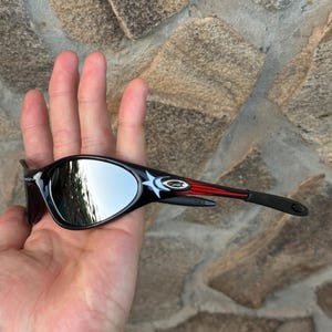 May include: Black sunglasses with mirrored lenses. The frames are black with red and white accents on the temples. The sunglasses are held in a hand against a stone background.