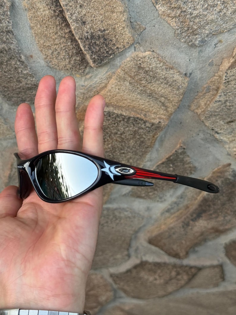 May include: Black sunglasses with mirrored lenses. The frames are black with red and white accents on the temples. The sunglasses are held in a hand against a stone background.
