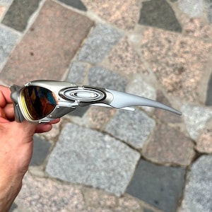 以下が含まれることがあります： 曲線的なデザインのシルバーとグレーのサングラス。レンズはオレンジと黄色のグラデーションになっています。フレームの側面にはOakleyのロゴがあります。サングラスは石の背景に対して保持されています。