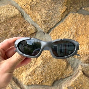 May include: Gray sunglasses with a unique, angular design. The lenses are dark, reflecting the surroundings. The frames have a textured, metallic appearance, with small decorative details near the temples. The sunglasses are held against a stone background.