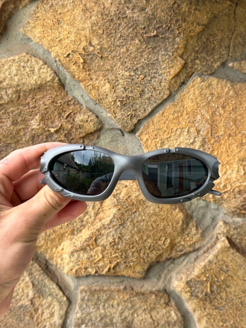 May include: Gray sunglasses with a unique, angular design. The lenses are dark, reflecting the surroundings. The frames have a textured, metallic appearance, with small decorative details near the temples. The sunglasses are held against a stone background.