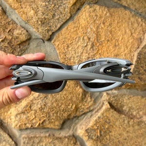 May include: Gray sunglasses with a futuristic design. The sunglasses have a silver frame and dark lenses. The arms of the sunglasses are angular and extend outward. The sunglasses are held against a stone background.