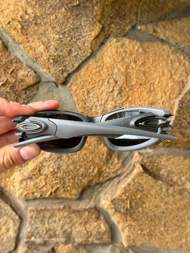 May include: Gray sunglasses with a futuristic design. The sunglasses have a silver frame and dark lenses. The arms of the sunglasses are angular and extend outward. The sunglasses are held against a stone background.