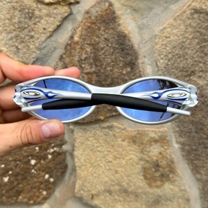 May include: White and silver sunglasses with blue lenses. The sunglasses have a black bridge and the brand logo on the temples. The frames have a flame design in blue. The sunglasses are held against a stone background.