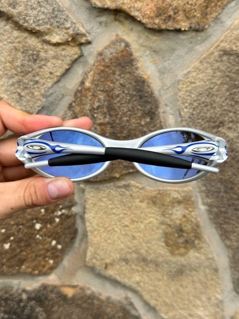 May include: White and silver sunglasses with blue lenses. The sunglasses have a black bridge and the brand logo on the temples. The frames have a flame design in blue. The sunglasses are held against a stone background.