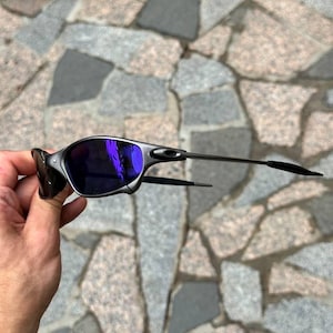 May include: A pair of sunglasses with a silver frame and dark purple lenses. The sunglasses have a sleek, modern design with thin temples. The lenses reflect a bright purple hue. The sunglasses are held by a hand.