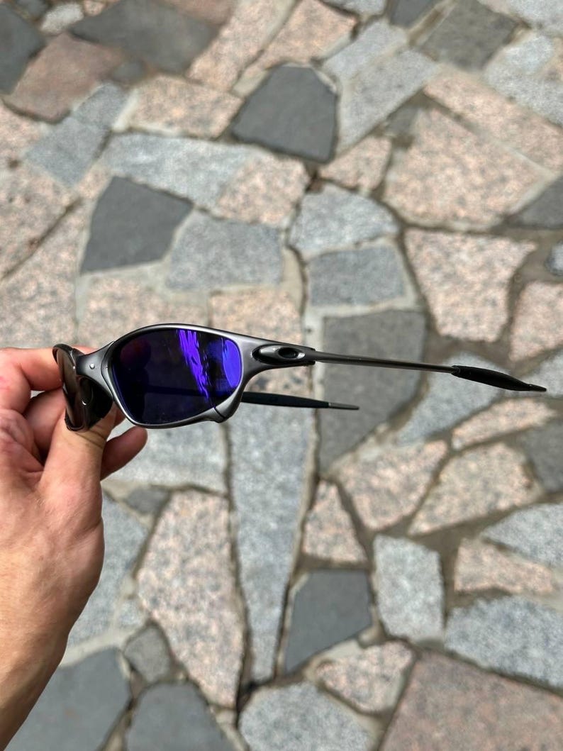 May include: A pair of sunglasses with a silver frame and dark purple lenses. The sunglasses have a sleek, modern design with thin temples. The lenses reflect a bright purple hue. The sunglasses are held by a hand.