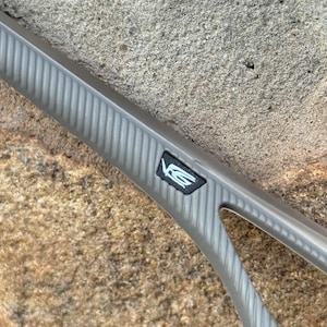 May include: A close-up of a gray automotive part with a textured, ribbed surface. A black emblem with white lettering is visible. The part is set against a rough, tan stone background.
