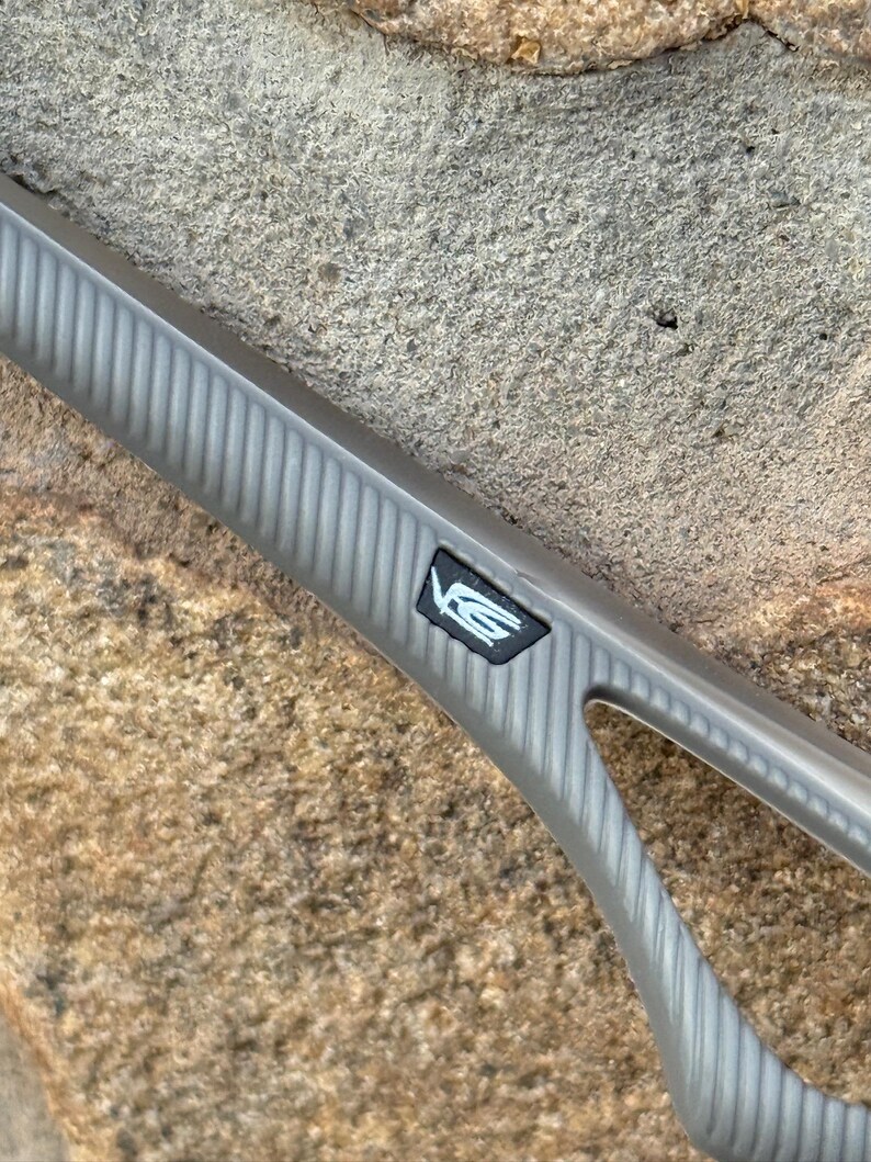 May include: A close-up of a gray automotive part with a textured, ribbed surface. A black emblem with white lettering is visible. The part is set against a rough, tan stone background.