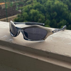 May include: A pair of sporty sunglasses with a silver and black frame and dark lenses. The sunglasses are resting on a white wooden surface, with a green, leafy background.