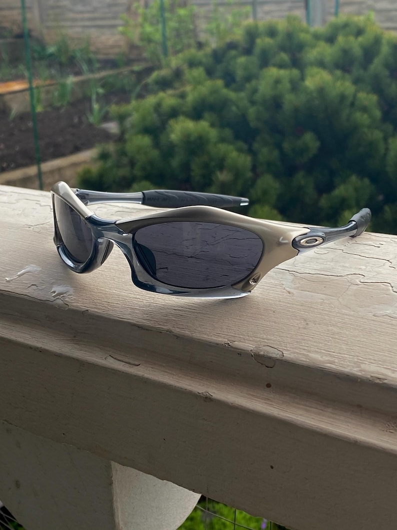 May include: A pair of sporty sunglasses with a silver and black frame and dark lenses. The sunglasses are resting on a white wooden surface, with a green, leafy background.